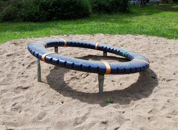 Auf einem Sandplatz, umgeben von Wiese, ist ein schräger blau-orangener Ring.