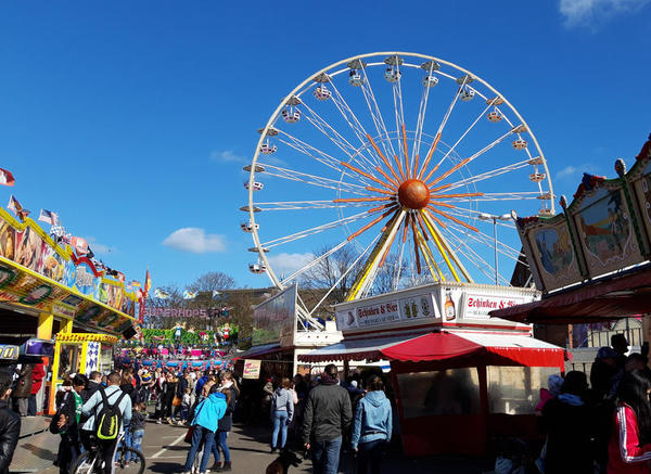 Besucher*innen vergnügen sich  auf dem Jahrmarkt. Ein Riesenrad ragt in den blauen Himmel.