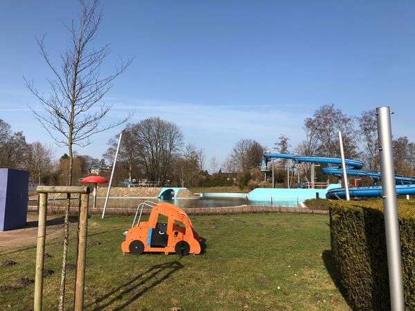 Im Bild sind im Vordergrund ein Kinderspielplatz und im Hintergrund das große Freibad mit blauer Rutsche zu sehen.