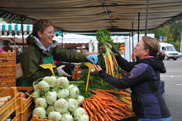 Eine Frau kauft am Gemüsestand ein und hält ein Bund bunte Möhren in der Hand, während die Marktfrau ihr das Wechselgeld zurückgibt.