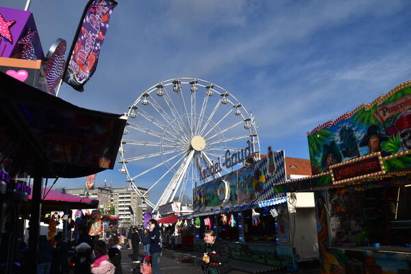 Viele Besucherinnen und Besucher des Jahrmarktes zwischen Jahrmarktbuden. Im Hintergrund steht ein sehr großes Riesenrad.