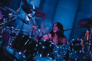 Geschminkte Schlagzeugerin spielt lächelnd Schlagzeug.