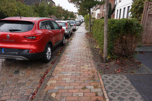 Das Foto zeigt den mit Pflastersteinen verlegten Gehweg. Links stehen geparkte Autos, rechts sind eine grüne Hecke und Bäume.