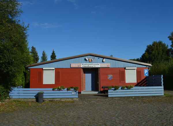 Das rote Vereinshaus für die Sportler*innen ist aus Holz und hat einen blauen Zaun.