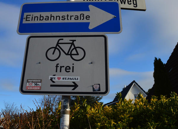 Unter dem Verkehrsschild für die Einbahnstraße zeigt ein weiteres die freie Nutzung für Fahrräder an. Das Schild ist Aufklebern beklebt.