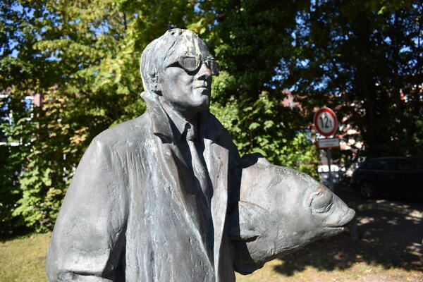 Die Bronzefigur mit den markanten Gesichtszügen trägt eine Sonnenbrill und einen Fisch unter dem Arm.