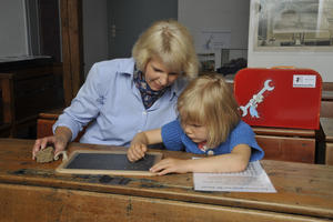 Ein junges Besucherkind übt auf einer kleinen Schiefertafel das Schreiben der altdeutschen Schrift in einem historischen Klassenzimmer.