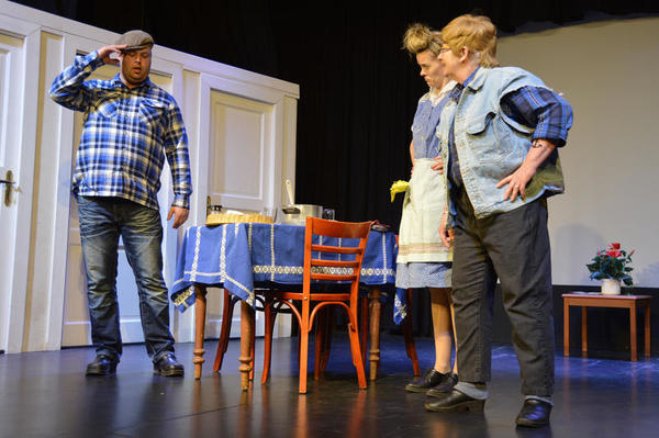 Eine Szene auf der Theaterbühne zeigt drei Akteure an einem Tisch.