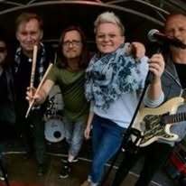 Vier Personen mit verschiedenen Musikinstrumenten und Mikrofon stehen auf einer B&uuml;hne und l&auml;cheln in die Kamera.