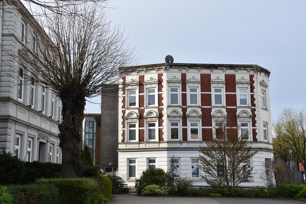 Links im Bild ein wei&szlig;es Mehrfamilienhaus mit einem Baum davor. Daneben ein weiteres Mehrfamilienhaus mit roten Klinkersteinen und wei&szlig;en Fenstern auf einem wei&szlig;en Sockel