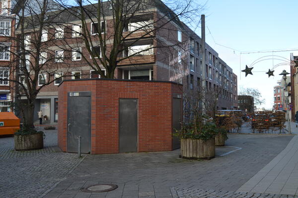 Bild vergr&ouml;&szlig;ern: Eine sechseckige WC-Anlage aus rotem Backstein mit Metallt&uuml;ren, wovon eine T&uuml;r den Zugang f&uuml;r Menschen mit Behinderung darstellt.