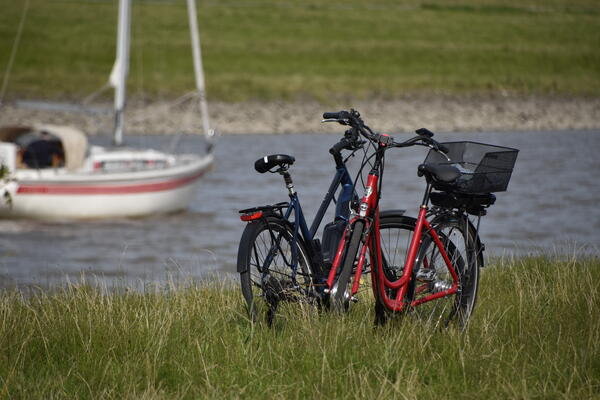 Zwei Fahrräder stehen im Gras am Ufer der Krückau. Ein kleines Segelschiff fährt vorbei.