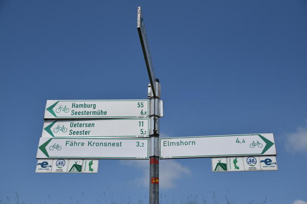 Ein Wegweiser zeigt die Entfernung und die Richtung für verschiedene Fahrrad Routen an. Er steht vor einem blauen Himmel.