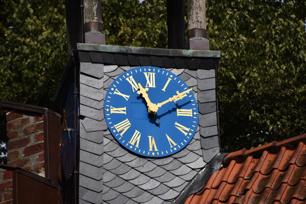 Die blaue Kirchturmuhr der historischen Kirche in Seester zeigen acht Uhr an. Das große Ziffernblatt hat goldfarbene, römische Ziffern und Zeiger. Sie leuchten in der Sonne.