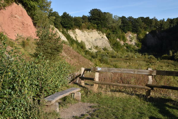 Eine Holzbank aus Bohlen befindet sich vor dem Weg in die Schlucht. An den Gesteinsformationen sind verschiedene geologische Gesteinsschichten sichtbar. 