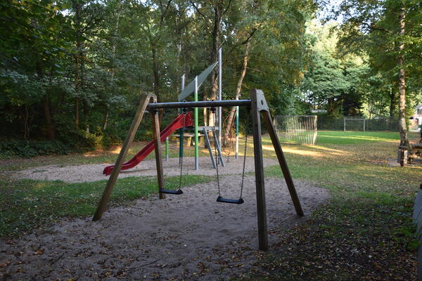 Eine Schaukel und eine Rutsche stehen auf dem kleinen Spielplatz unter Bäumen.