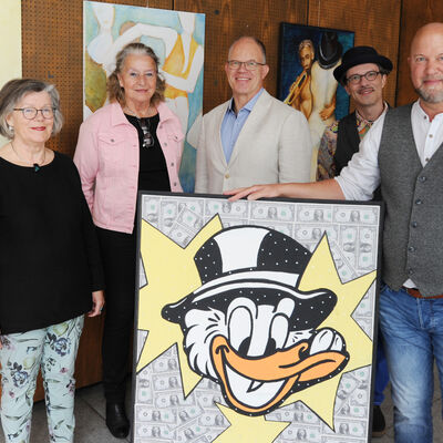 Das Foto zeigt zwei Frauen und drei M&auml;nnervor einer Wand, an der drei Gem&auml;lde zu sehen sind. Ein weiteres gro&szlig;formatiges Bild mit Dagobert im Mittelpunkt steht vor der Menschengruppe im Vordergrund.