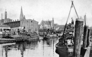 Bild vergr&ouml;&szlig;ern: Das Bild zeigt eine Schwarzwei&szlig;-Aufnahme des Elmshorner Hafens mit Schiffen auf dem Wasser und Geb&auml;uden im Hintergrund.
