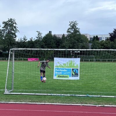 Das Foto zeigt einen Fu&szlig;ballspieler auf dem Sportplatz, der einen Ball ins leere Tor schie&szlig;t. Im Netzt des Tores ist ein Plakat aufgeh&auml;ngt, auf dem der Slogan Fairtrade-Stadt Elmshorn geschrieben steht.