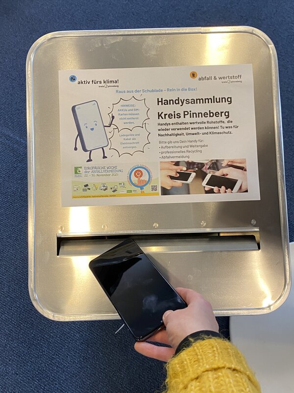 Eine Hand wirft ein Handy in eine silberne geschlossene Alu-Sammelbox.