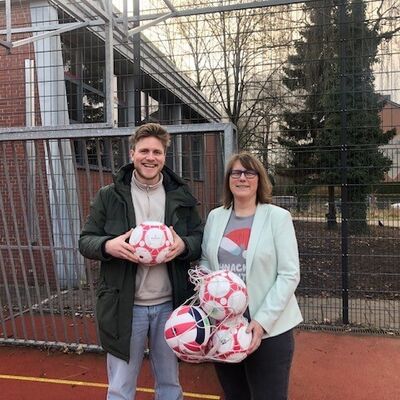 Eine Frau und ein Mann halten B&auml;lle in der Hand, die sich zum Teil in einem Ballnetz befinden. Die beiden Personen befinden sich auf einem Sportgel&auml;nde, das von einem Metallzaun eingegrenzt ist.