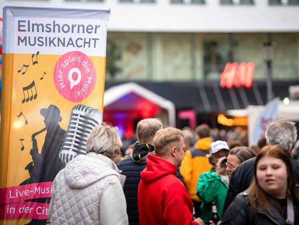 Links im Bild eine Beachflag mit der Aufschrift Elmshorner Musiknacht. Weiter ist auf dem Banner eine Person in schwarzer Kontur abgebildet, die Saxophon spielt. Neben dem Banner befinden sich viele Menschen im Geschehen.