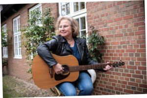 Frau sitzt mit einer Gitarre vor einer Hauswand.