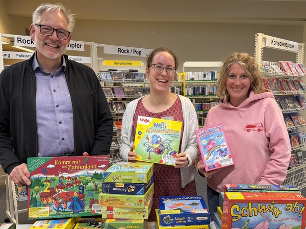 Zwei Frauen und ein Mann mit Gesellschaftsspielen vor gef&uuml;llten B&uuml;cherregalen.