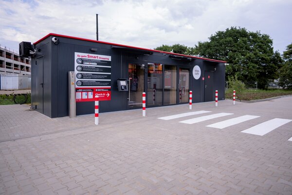 Bild vergr&ouml;&szlig;ern: Ein Smart Shop untergebracht in einem Container &auml;hnlichen Geb&auml;ude -  davor Markierungspfosten und ein Zebrastreifen f&uuml;r die sichere Zuf&uuml;hrung zum Eingang.