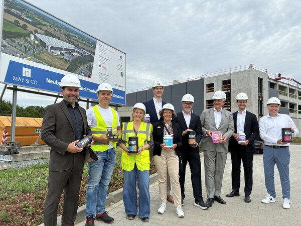 Frauen und M&auml;nner - darunter Elmshorns Oberb&uuml;rgermeister Erik Sachse und Zweiter Stadtrat Marius Munk - vor einer Gro&szlig;baustelle eines Unternehmens, Im Hintergrund sind Teile des Geb&auml;udes und ein &uuml;berdimensionales Bauschild zu sehen.