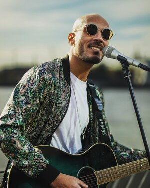 Bild vergr&ouml;&szlig;ern: Musiker Shebab mit Sonnenbrille, lockerem Hemd und Gitarre singt open Air in ein Mikrofon.