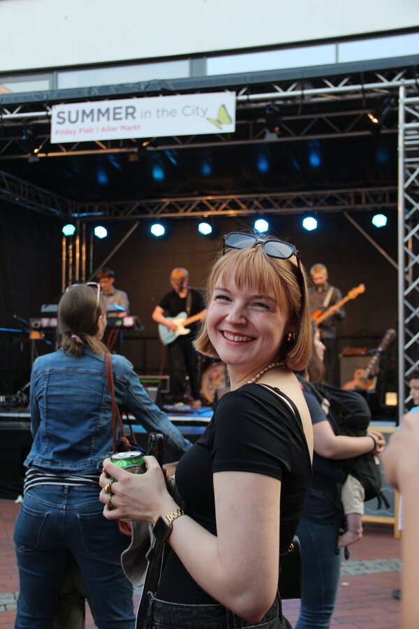 Eine Frau guckt sich vor einer Musikb&uuml;hne mit spielender Band zum Fotografen um.