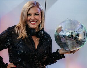 Bild vergr&ouml;&szlig;ern: Eine blonde Frau im Glitzerkleid und mit Musikkopfh&ouml;rer um den Hals h&auml;lt eine Discokugel in der Hand.
