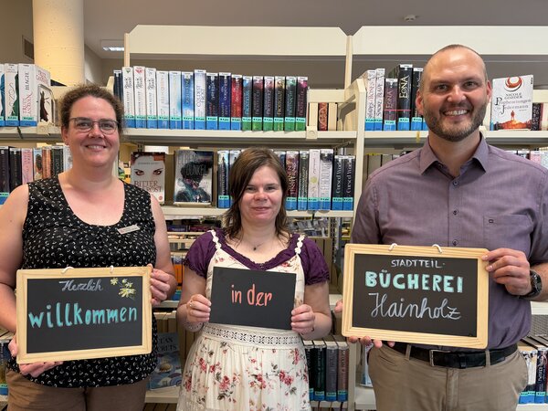 Zwei Frauen und ein Mann stehen vor B&uuml;cherregalen. In den H&auml;nden halten sie Tafeln auf denen steht: "Herzlich willkommen in der Stadtteil-B&uuml;cherei Hainholz".