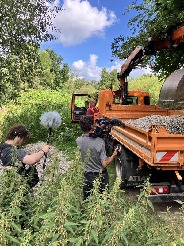Bild vergr&ouml;&szlig;ern: Ein Filmteam des NDR bei Dreharbeiten am Kr&uuml;ckau-Ufer vor einem Lkw mit Kies auf der Ladefl&auml;che und einer Baggerschaufel.
