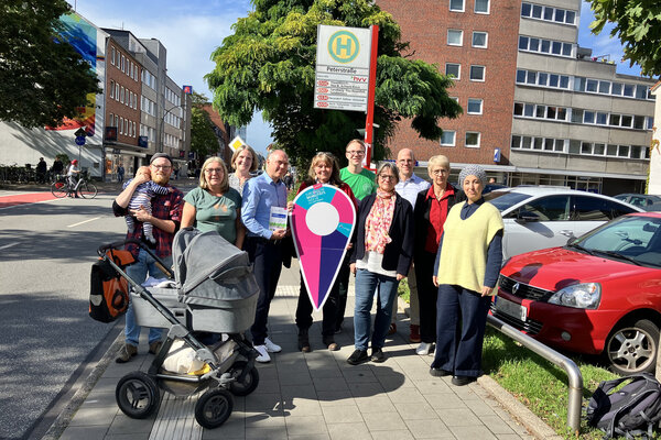 Bild vergr&ouml;&szlig;ern: Eine Gruppe von Personen, die auf dem rechten B&uuml;rgersteig der Schulstra&szlig;e in Elmshorn in H&ouml;he des Parkplatzes in Nachbarschaft zur Wei&szlig;en Villa an einer Bushaltestelle steht und f&uuml;r Europ&auml;ische Mobilit&auml;tswoche 2026 wirbt.