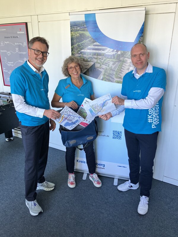 Die drei Personen stehen in blauen T-Shirts mit dem Aufdruck #Kreis Pinneberg Wirtschaft vor einer Wand und packen eine blaue Tasche.