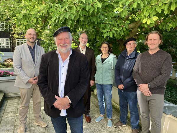Bild vergr&ouml;&szlig;ern: Sechs Personen stehen auf einer gepflasterten Fl&auml;che vor einem gr&uuml;n belaubten Baum. Im Hintergrund ist ein rot geklinkertes Geb&auml;ude mit wei&szlig;en Fenstern zu sehen.