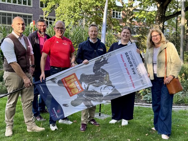 Bild vergr&ouml;&szlig;ern: Sechs Frauen und M&auml;nner auf dem gr&uuml;nen Rasen mit einer Werbeflagge in den H&auml;nden.