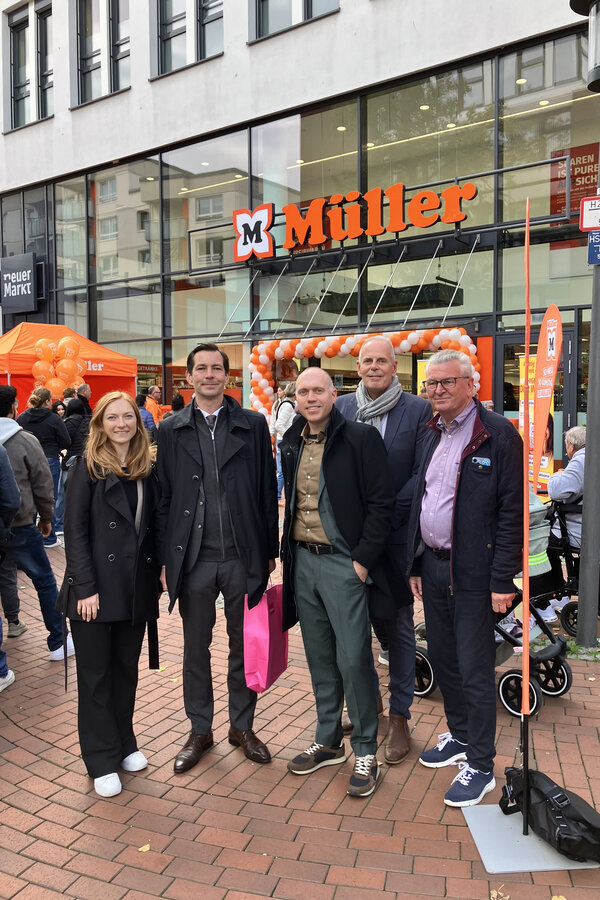Bild vergr&ouml;&szlig;ern: F&uuml;nf Personen vor einer orange-wei&szlig; geschm&uuml;ckten M&uuml;ller-Filiale. Drum herum str&ouml;men Leute in die und aus der Filiale.