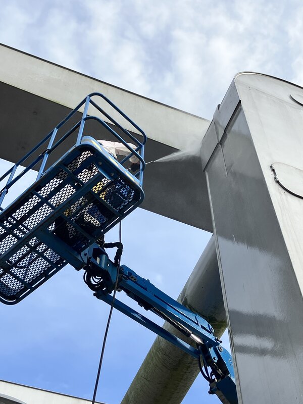 Bild vergr&ouml;&szlig;ern: Ein Mann in einem blauen Hubsteiger-Korb strahlt Metallteile der Br&uuml;cke mit einem Hochdruckreiniger ab.