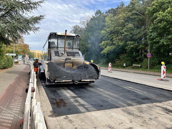 Bild vergr&ouml;&szlig;ern: Ein Baufahrzeug f&auml;hrt auf einer abgefr&auml;sten Stra&szlig;e und verteilt neuen Asphalt darauf.