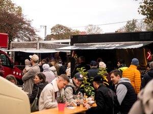 Bild vergr&ouml;&szlig;ern: Viele Menschen sitzen essend zwischen den Food Trucks am Tisch zusammen.