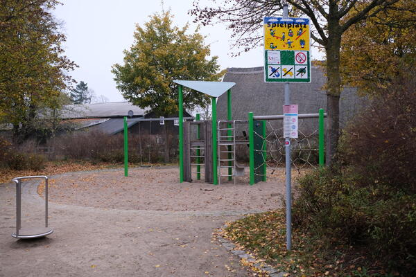 Grün gestrichenes Metall-Klettergerüst auf dem Spielplatz Paul-Klee-Straße. Im Hintergrund ein großer Baum neben einem alten Bauernhaus.