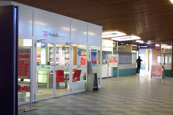 Innenansicht des Bahnhofgebäudes mit Blick auf die gläserne Eingangstür und das Schaufenster des Reisezentrums. Es ist hell und einladend,