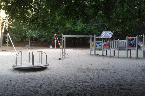 Auf einem Sandplatz umgeben von Bäumen stehen verschieden Spielplatzgeräte: eine Drehscheibe, eine Reifenschaukel und ein Spielhaus.