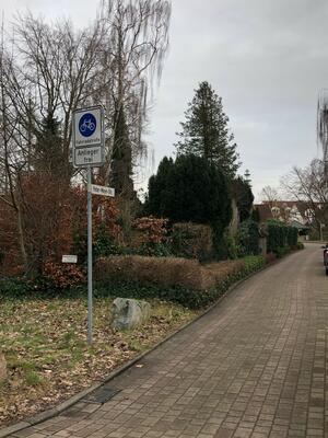 Ein Verkehrsschild mit der Aufschrift  "Fahrradstraße" steht neben einem gepflasterten Weg, der von gepflegten Bäumen und Gehölzen gesäumt wird.