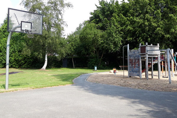 Auf einem großen Asphaltplatz mit Basketball-Korb ist eine Wiese und ein Sandplatz mit Klettergerüst aus Holz und Metalstangen.