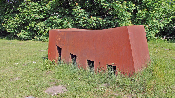 Das Kunstwerk des Künstlers Winni Schaak zeigt eine rostbraune Stahlkonstrukt mit eckigen Schächten auf einer Wiese im Skulpturengarten. Es trägt die Bezeichnung Architektur Eins uns ist aus dem Jahr 2000.