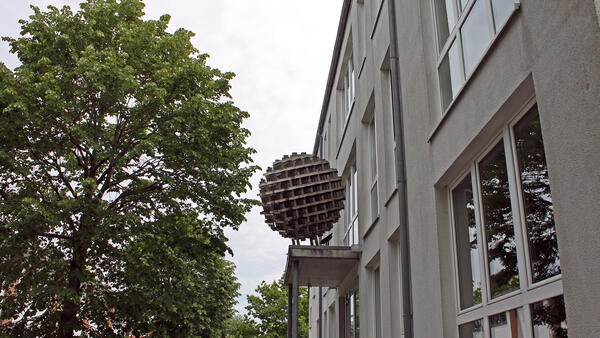 Eine massive Metallkugel des Künstlers HD Schrader mit der Bezeichnung Kugel aus den 1979er Jahren. Das Kunstwerk befindet sich auf einem Dachüberstand neben einer hellen Fassade in der Straße Langelohe 65.